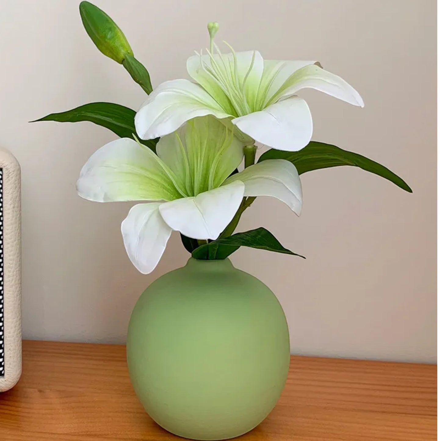 Sage-green bud vase with faux lilies next to a woven speaker on a bedside — Holm & Hearth