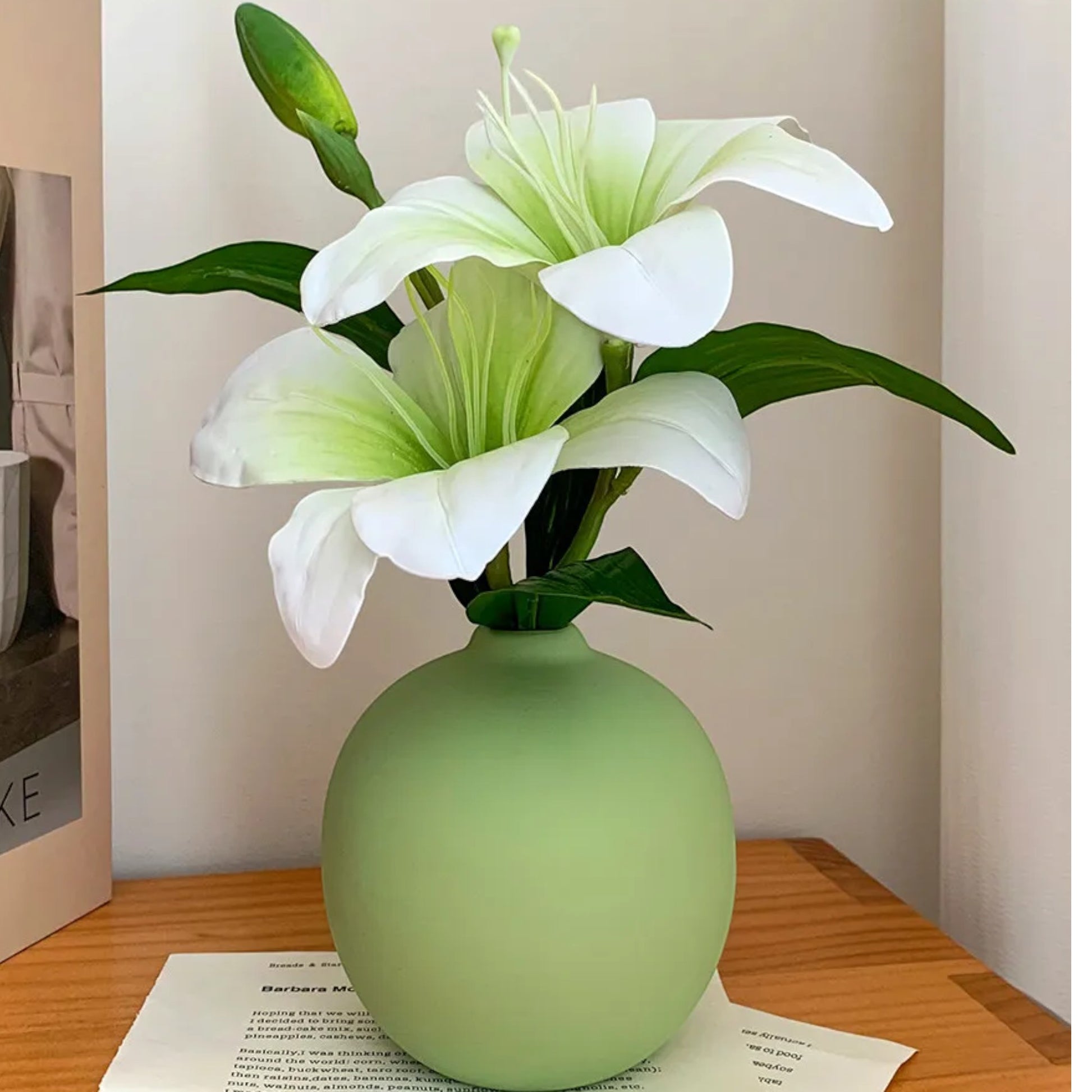 Sage-green ceramic bud vase with faux lilies on a wooden table, magazine behind — Holm & Hearth
