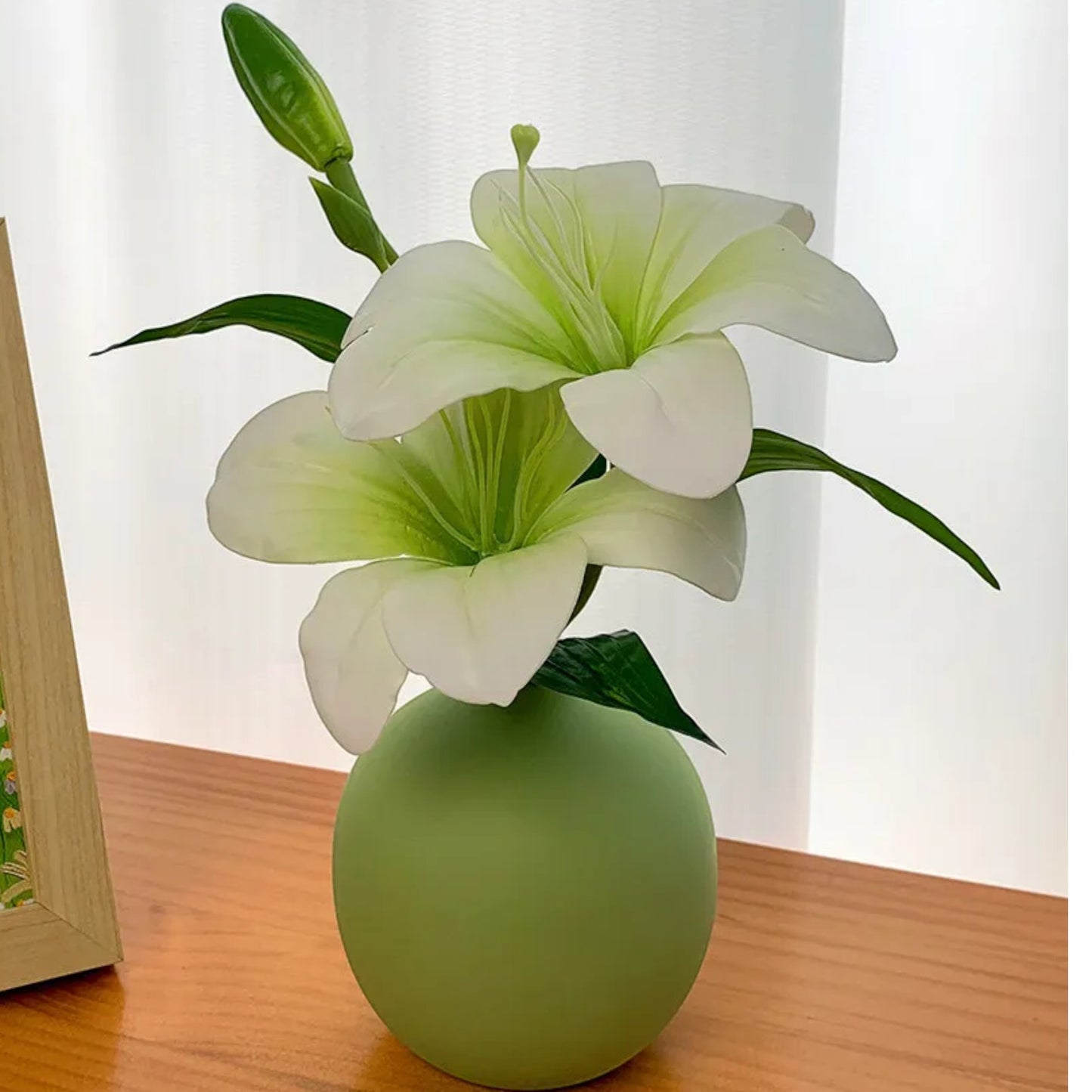 Single faux lily stem in a small green ceramic vase beside a framed painting — Holm & Hearth