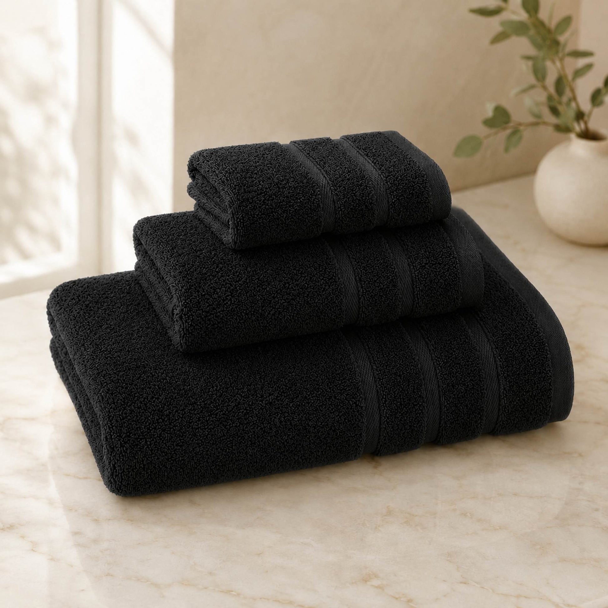 Pure cotton bath towel 3-piece set in black with satin edge, folded on warm cream marble surface, soft natural light