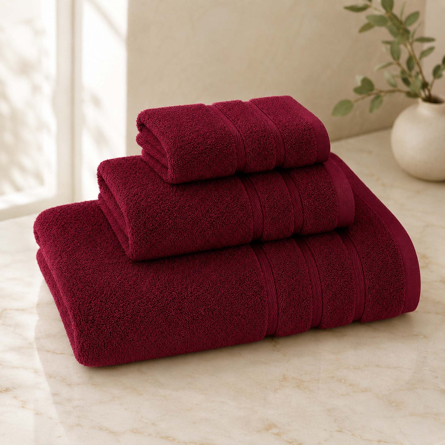 Pure cotton bath towel 3-piece set in burgundy with satin edge, folded on warm cream marble surface, soft natural light