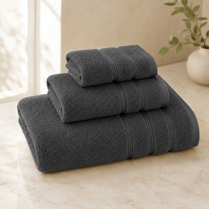 Pure cotton bath towel 3-piece set in charcoal with satin edge, folded on warm cream marble surface, soft natural light