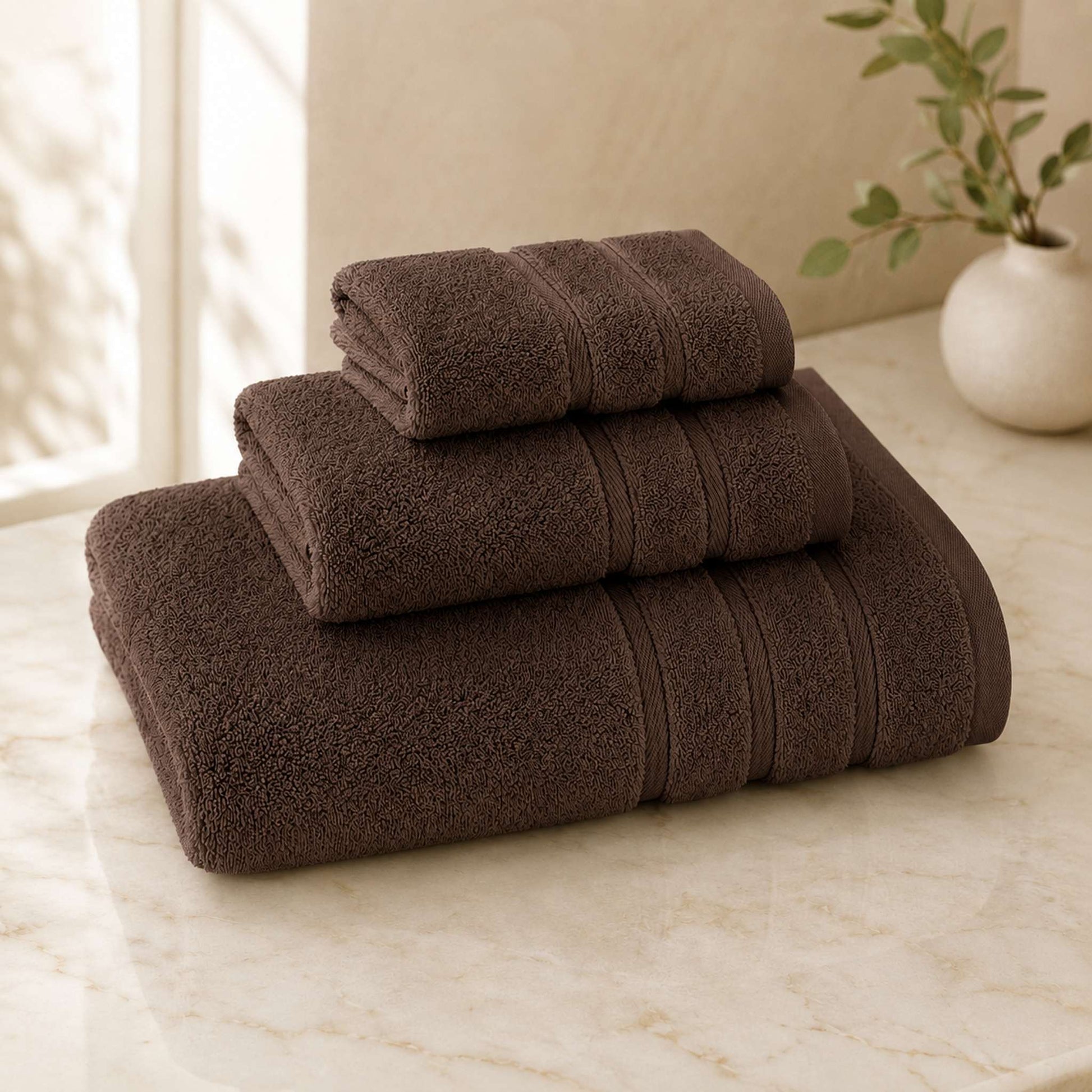 Pure cotton bath towel 3-piece set in cocoa with satin edge, folded on warm cream marble surface, soft natural light