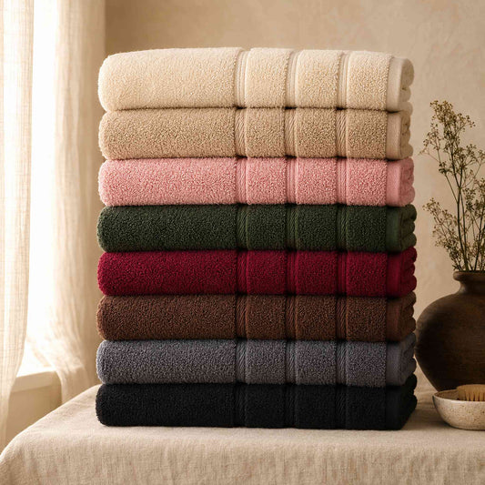 Pure cotton bath towel set in eight muted colours stacked on cream linen surface against soft taupe wall, hero shot