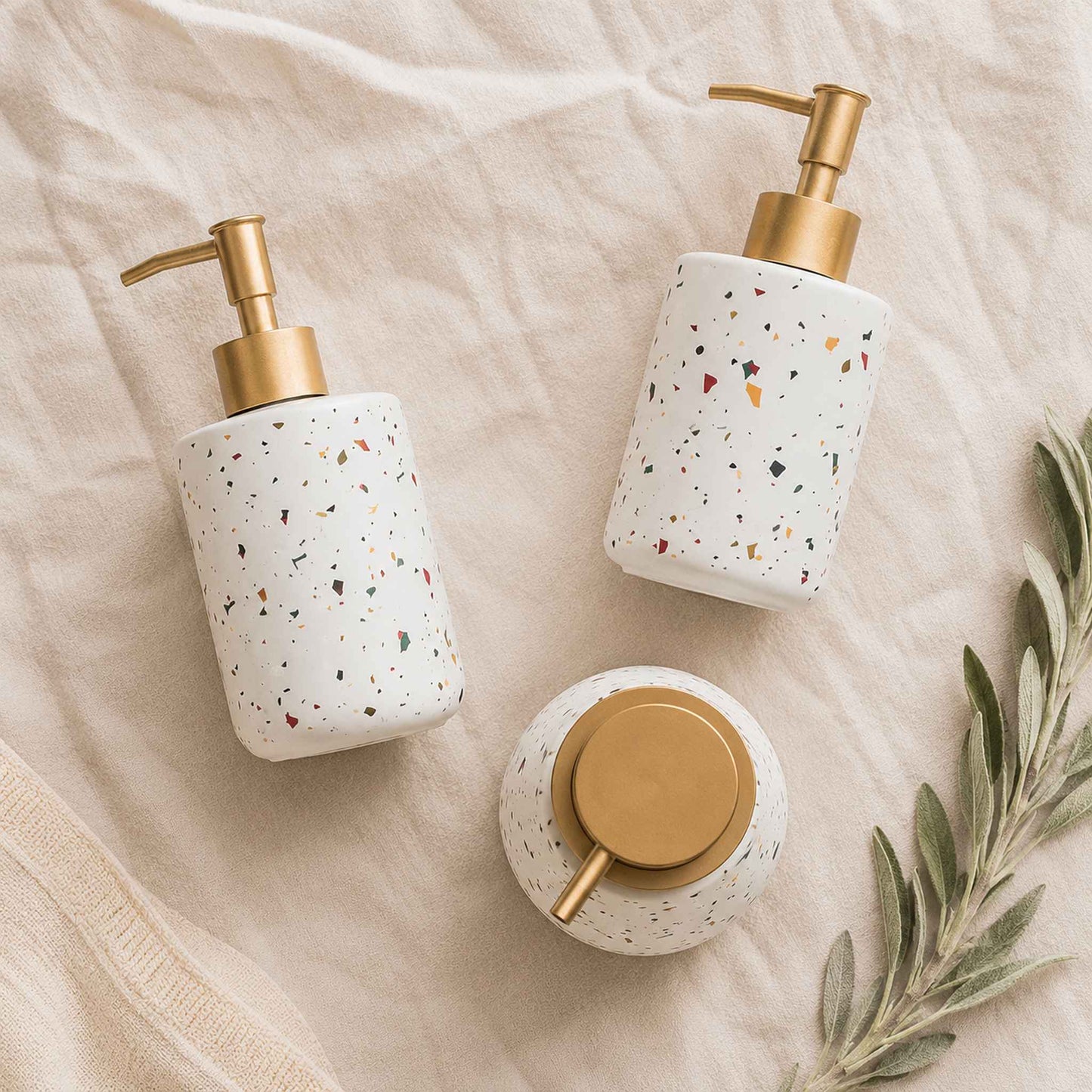 Three terrazzo ceramic soap dispenser bottles flat lay on warm off-white linen with dried sage sprig