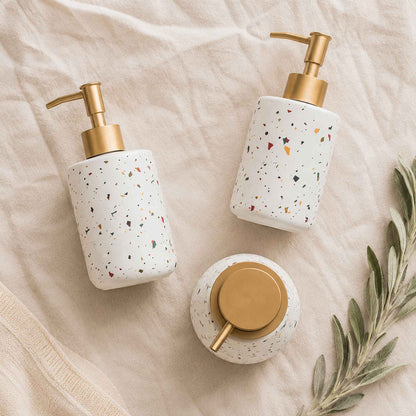 Three terrazzo ceramic soap dispenser bottles flat lay on warm off-white linen with dried sage sprig