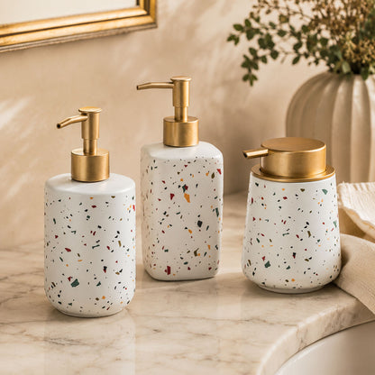 All three terrazzo ceramic soap dispenser bottles grouped on cream linen with brushed brass pumps, warm morning light