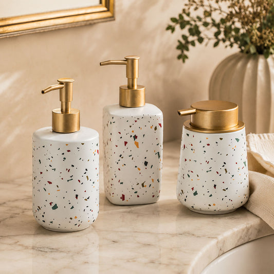All three terrazzo ceramic soap dispenser bottles grouped on cream linen with brushed brass pumps, warm morning light
