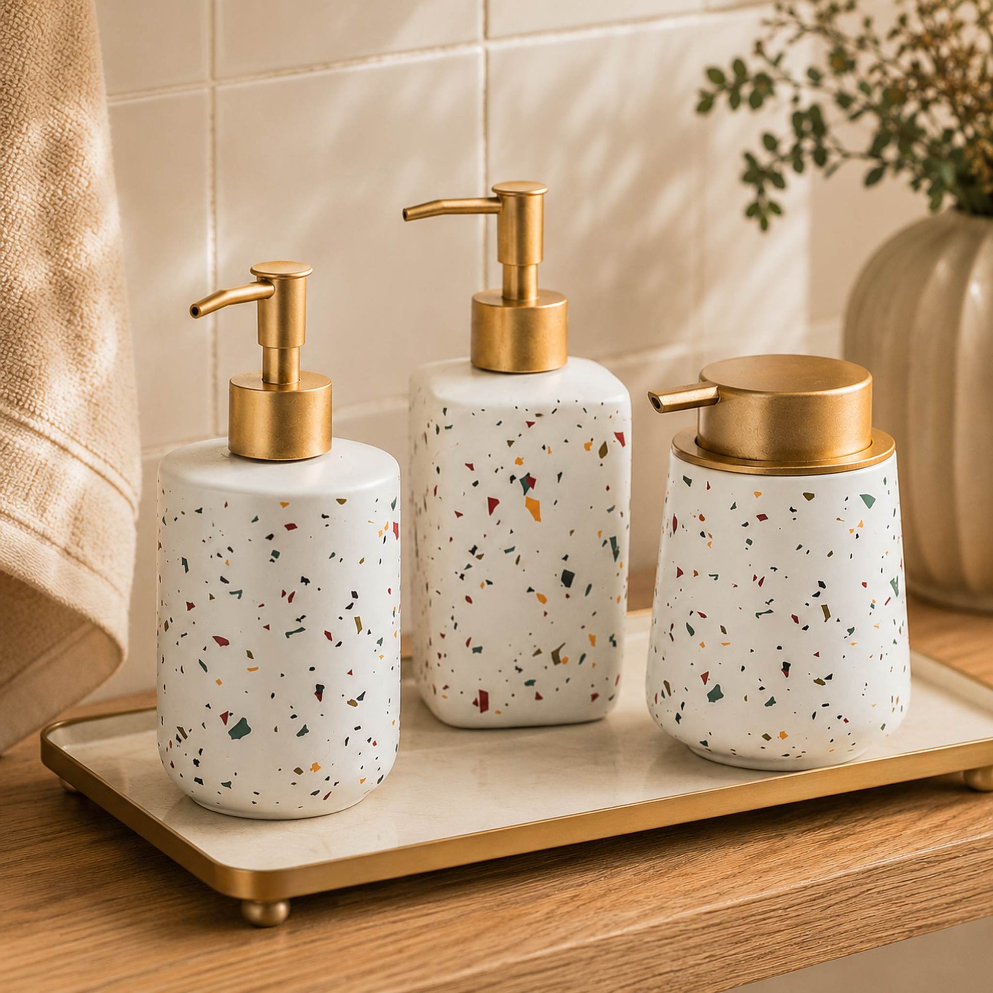 Three terrazzo ceramic soap dispenser bottles on brass-trimmed bathroom tray atop warm oak shelf, lifestyle shot