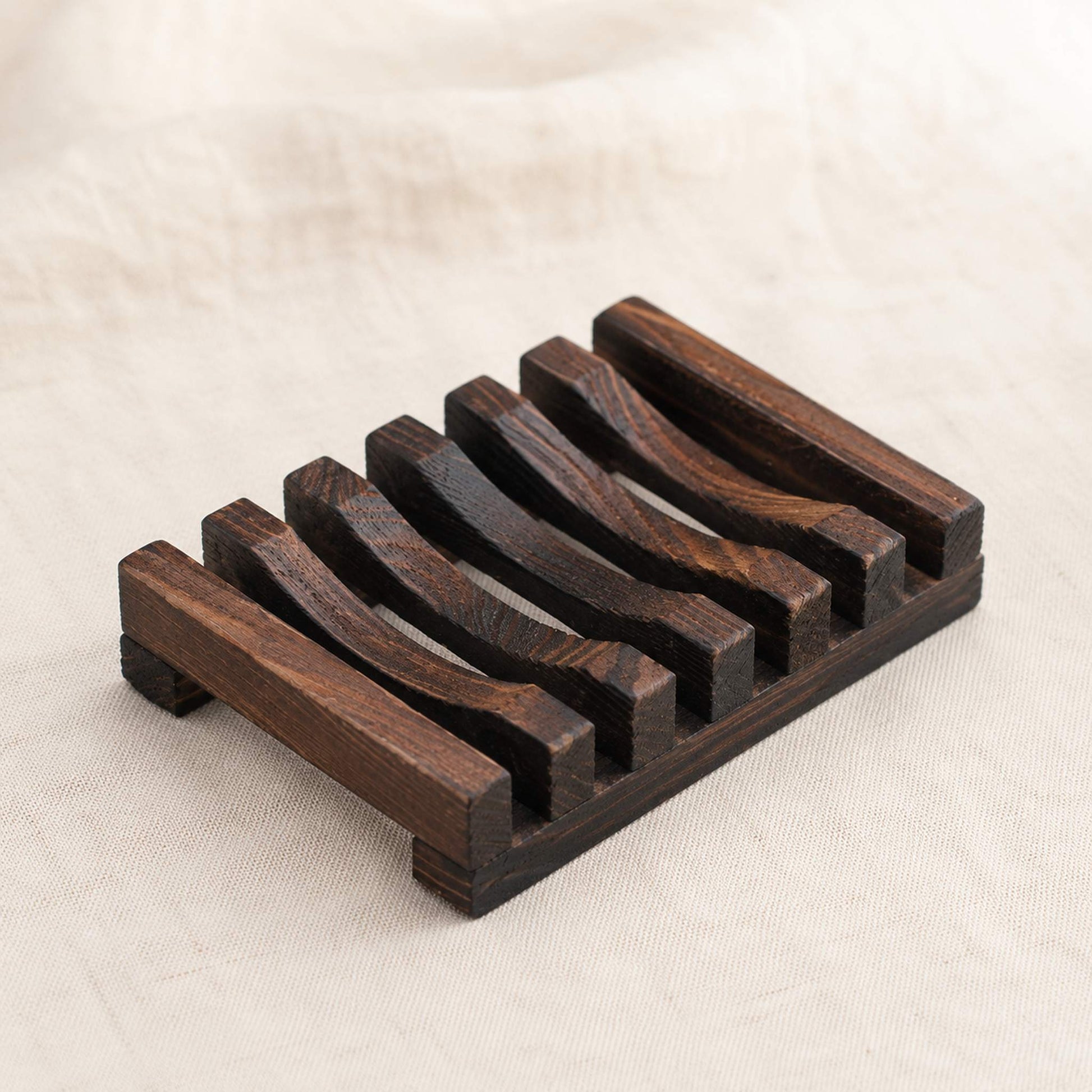 Charred wooden soap rack with drainage slats on cream linen surface, deep smoky finish with visible wood grain