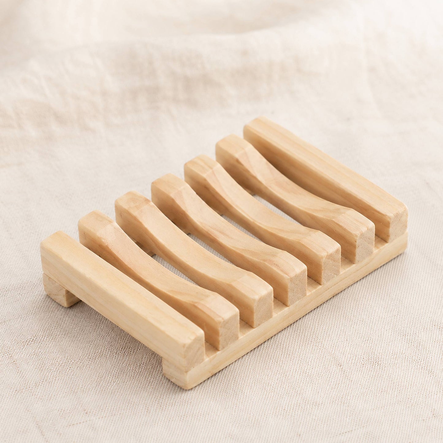 Natural wooden soap rack with drainage slats on cream linen surface, warm pale wood finish with visible grain